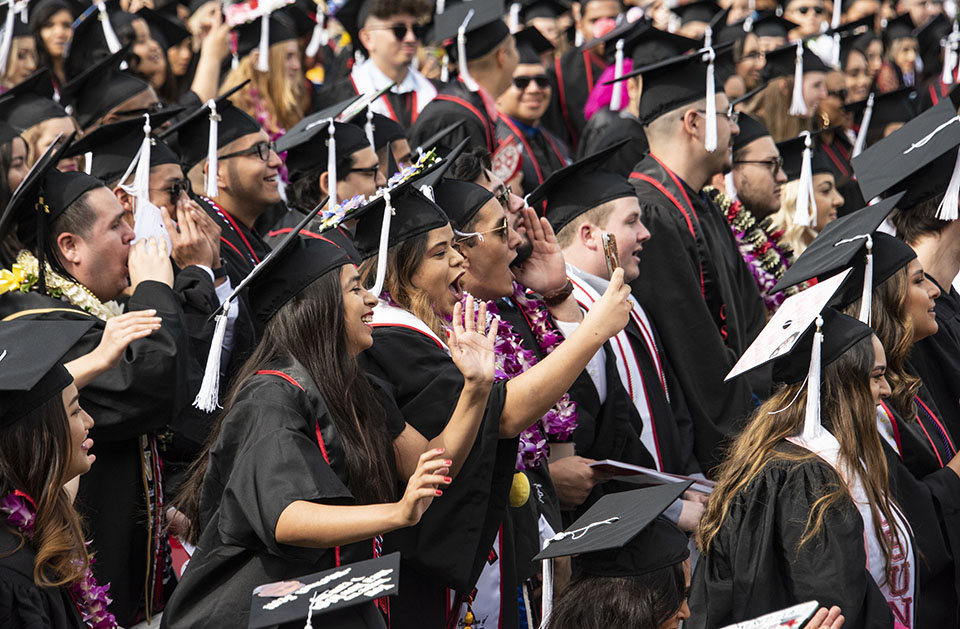 A crowd of college graduates smile and cheer during a graduation ceremony.