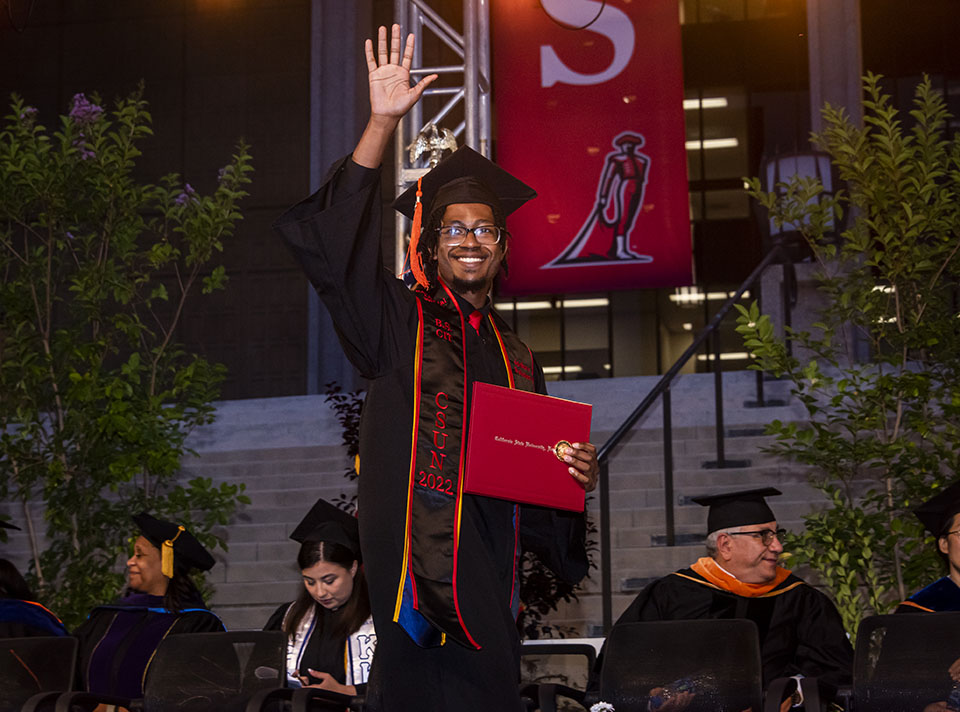 A new graduate in cap, gown and sash holds his red CSUN diploma case and waves as he crosses the Commencement stage.