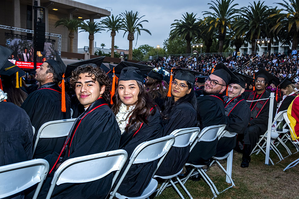 A crowd of CSUN graduates in caps, gowns and sashes, smile as they wait in their seats in front of the University Library, at Commencement 2022.