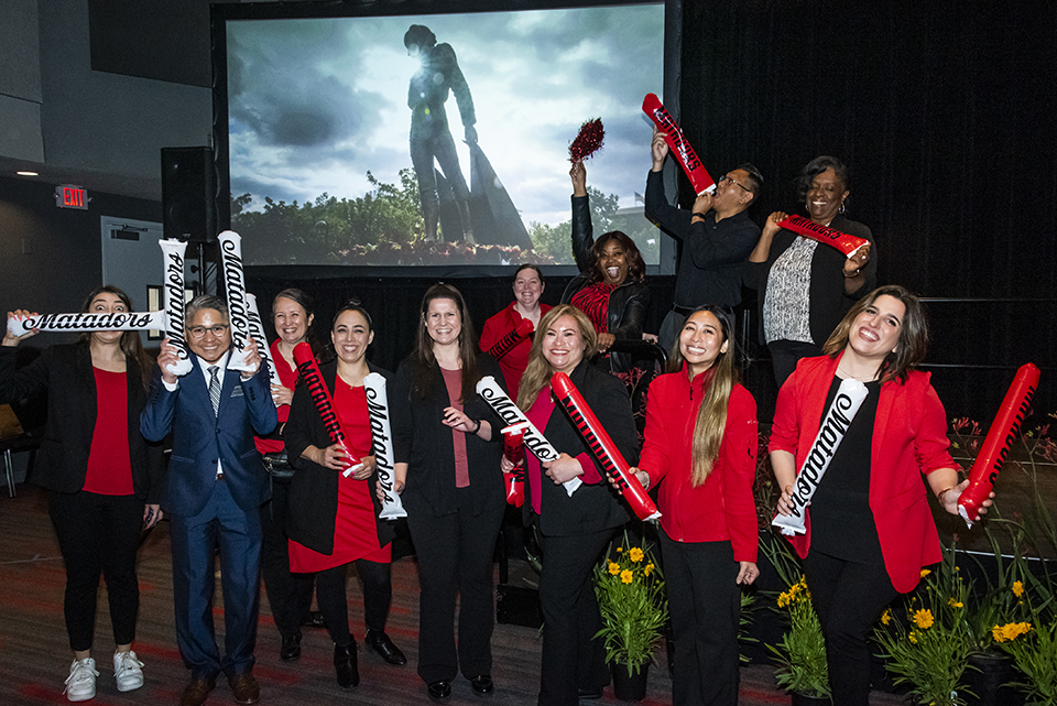 Staff members stand in front of a picture of the matador statue and hold red and white clappers that say 