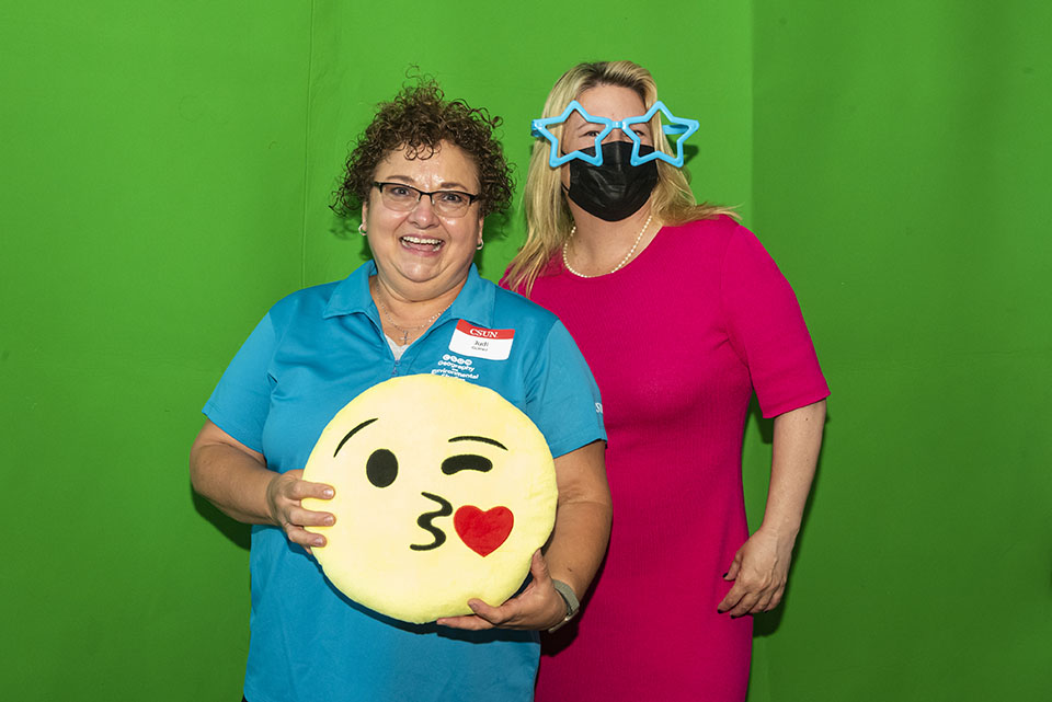 President Erika D. Beck is wearing large star-shaped glasses and is standing with another employee holding an emoji prop in front of a green screen in the photo booth.
