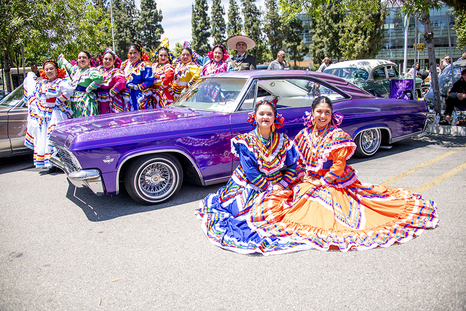 Dancers in brightly colored dresses are seated in front of and are standing behind a large purple sedan.