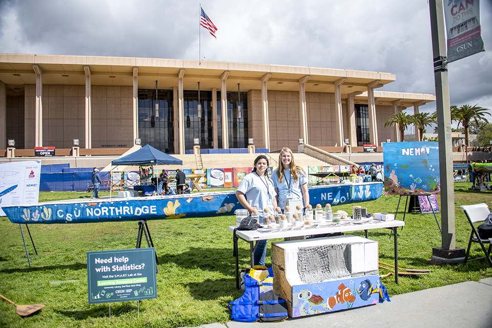two students stand on the University Library lawn in front of a blue-painted concrete canoe