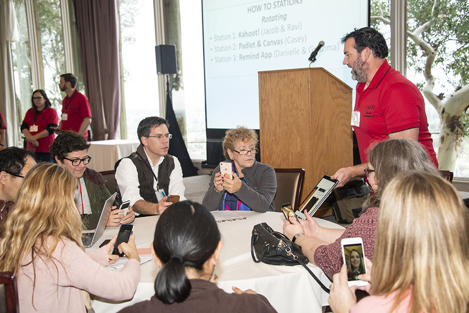 A man standing and demonstrating a program on a tablet, in front of a seated group of eight or nine CSUN faculty members.