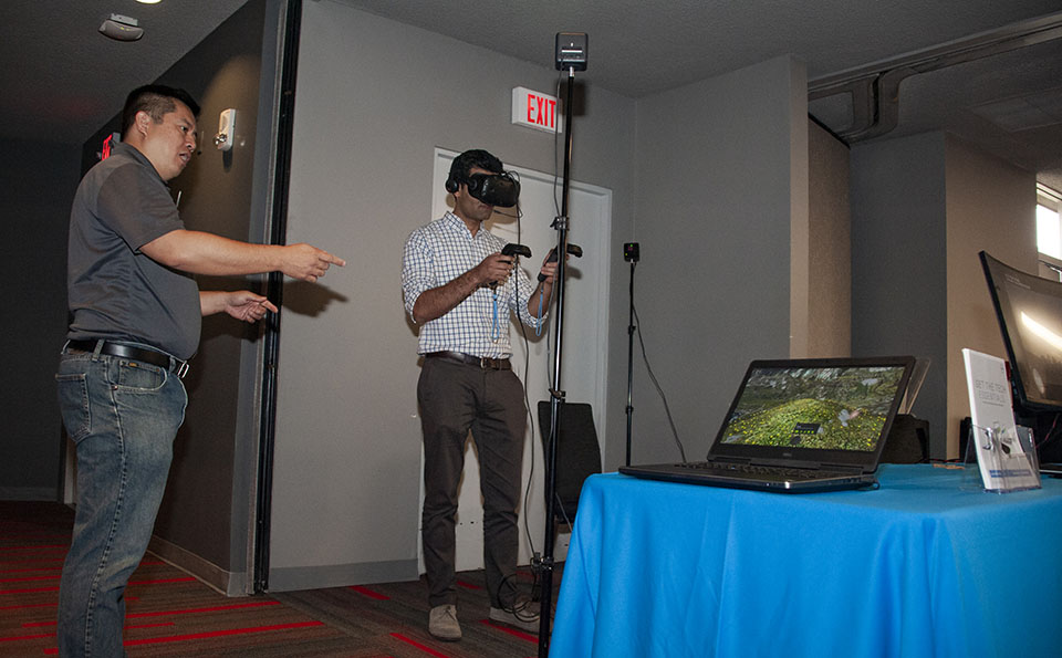 A man wears a virtual reality headset and holds two controllers, while a company representative points to a computer screen.