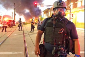 Man stands on street with fire behind him. He is wearing riot helmet and gas mask.