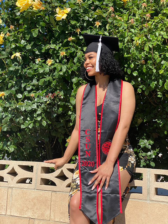 A CSUN student in a graduation cap and sash poses in a park setting.