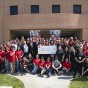 Students in red shirts surround faculty and staff for a group photo. At the center, administrators hold a large check.