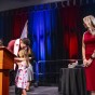 Landon Himes bends down to hug his young daughter on stage, while his wife stands off to the right and looks on.