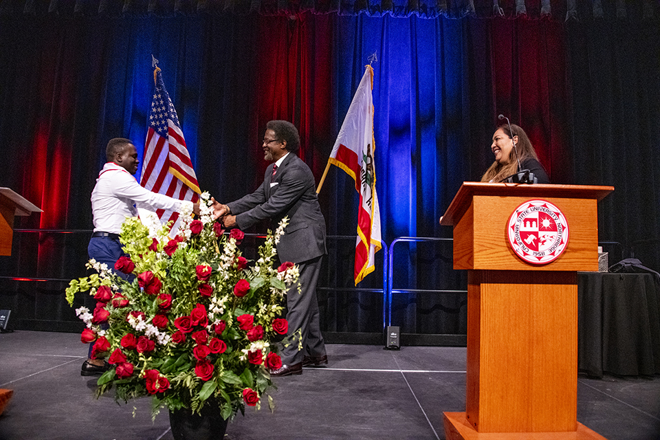 Kehinde Eijo shakes hands with William Watkins in the center of the stage while Mayra Plascencia looks on from the podium