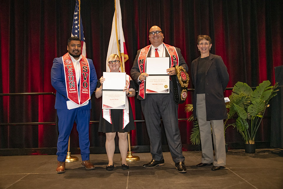 Four people stand on stage in front of flags. Two people in center show certificates