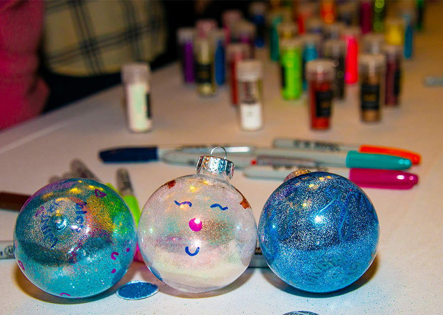 Three decorated Christmas tree ornaments photographed on the table in front of various art materials.