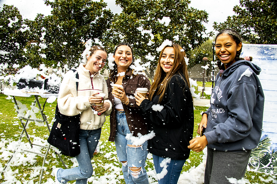 A group of four students covered in snow hold their cups of hot cocoa and smile for the photo.