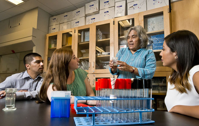 Biology professor MariaElena Zavala stands in a laboratory, showing several students a specimen in a test tube. A tray of test tubes sits on a table in front of her.