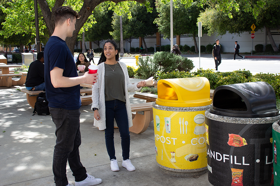 A CSUN student holds a reusable cup while another student disposes of compostable napkins in a compostable bin.