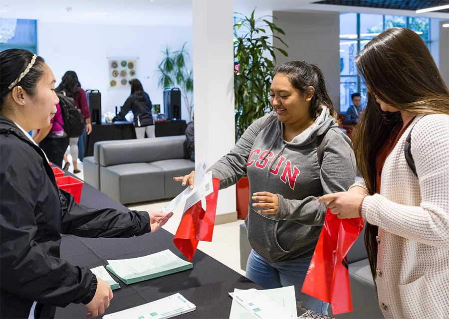 A student in a CSUN sweatshirt picks up Scantrwon sheets and a red tote bag, from a table in the USU.