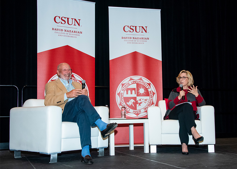 Wendy Greuel and alumnus Paul Dali sit in white chairs on the stage of CSUN's Northridge Center, with Nazarian College banners behind them. They are holding microphones, and Greuel is speaking.