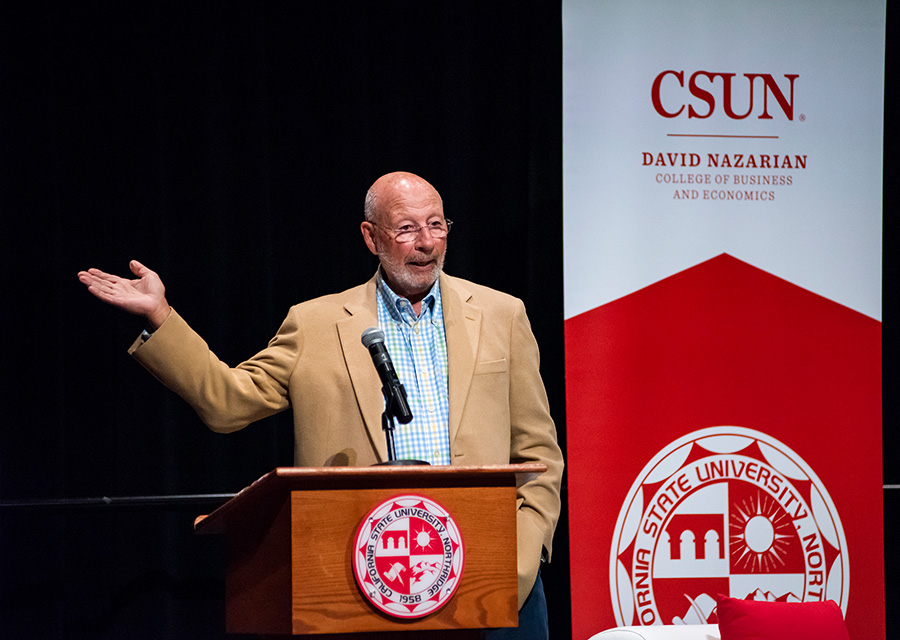 Tech and innovation legend and alumnus Paul Dali stands at a CSUN podium, next to a banner for the David Nazarian College of Business and Economics. He is speaking into a microphone and gesturing toward the audience.