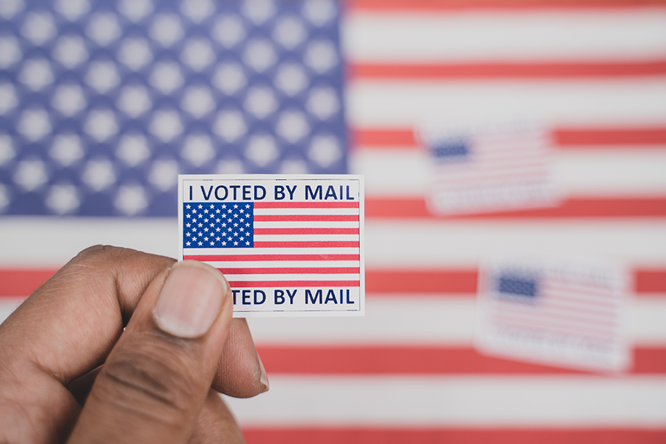 A hand holds an "I Voted by Mail" sticker against an American flag background.