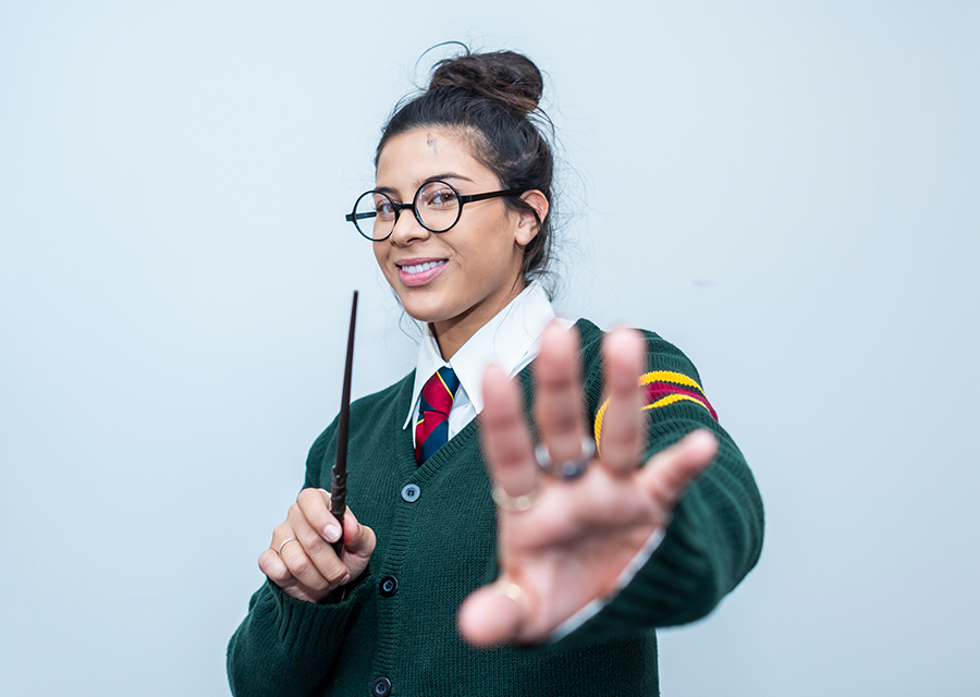 Karen Acevedo holds a hand towards the camera, in the other hand is a wand. Acevedo is wearing round glasses, a gray cardigan with red and gold stripes on the sleeve and a red tie. Acevedo has a lighting bolt drawn on her forehead