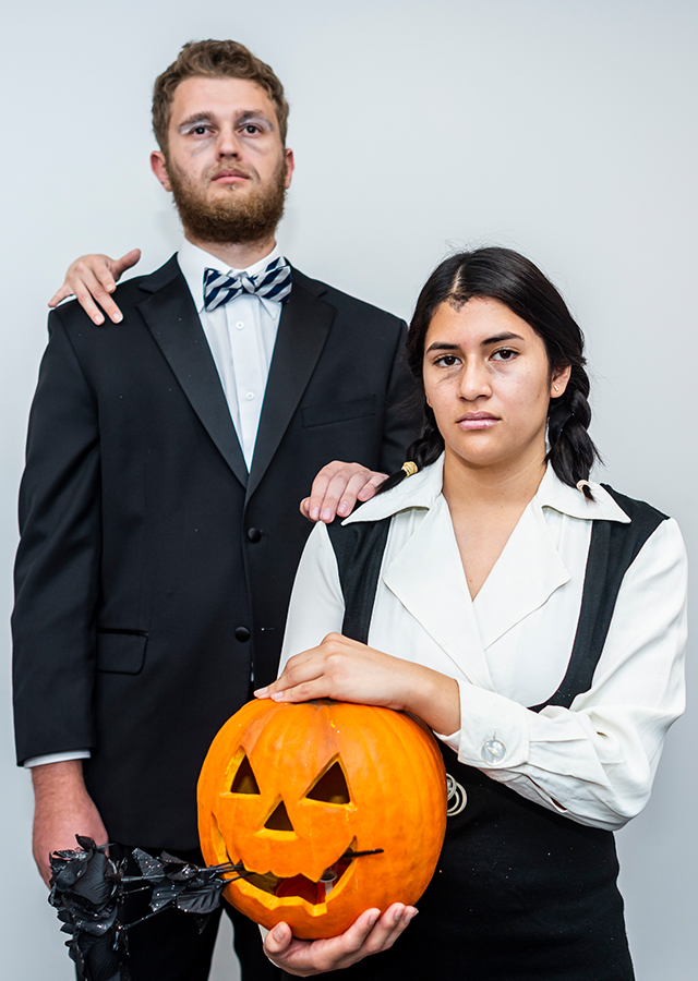 Ryan Silber and Michelle Hernandez stand side by side, glowering at the camera. Silber is wearing a suit and bowtie, with gray make up smudged under his eyes and on his face, and has a mysterious hand on his shoulder. Hernandez is wearing a black dress over a white collared shirt. Hernandez's hair is braided into pigtail and is hold a pumpkin in her hands.