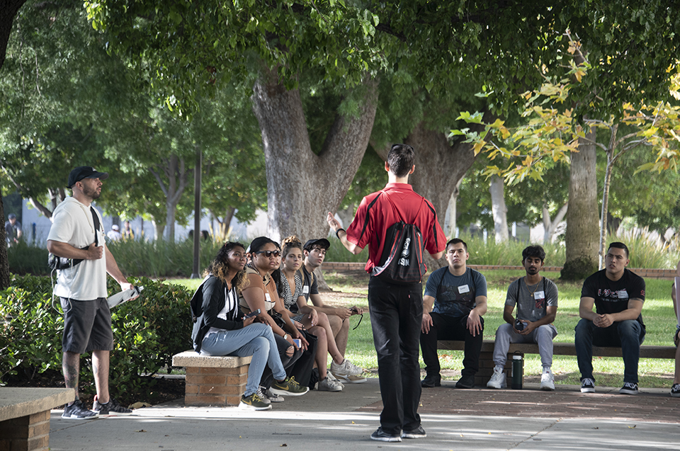 A group of incoming CSUN students sit on benches as they listen to a guide under a tall tree.