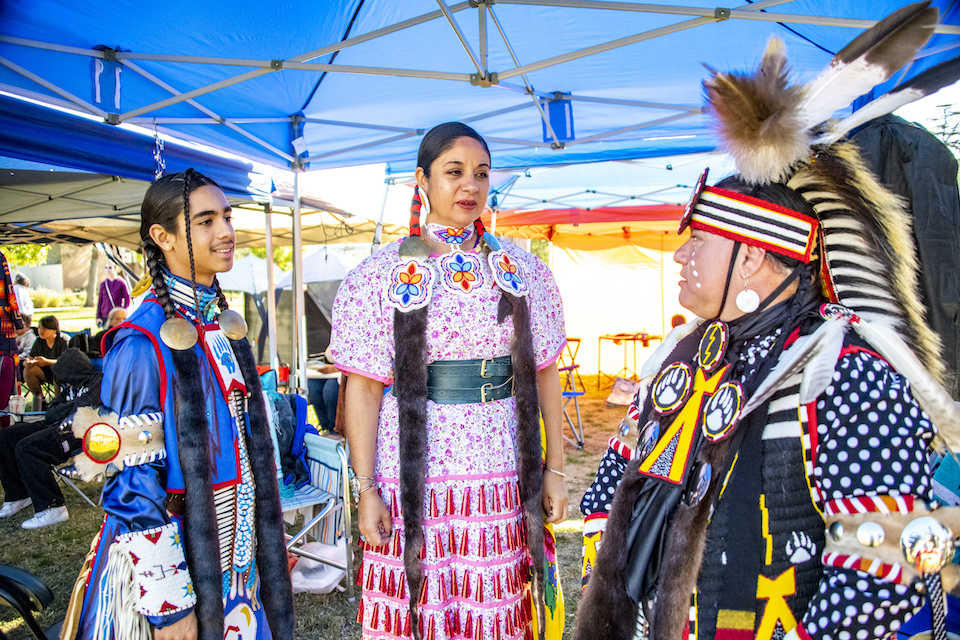 Three people stand in Native American regalia. Woman in center wears pink dress. Man on right wears feathered headdress