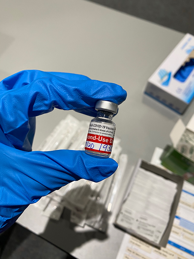 A blue gloved hand holds a vial of COVID-19 vaccine.